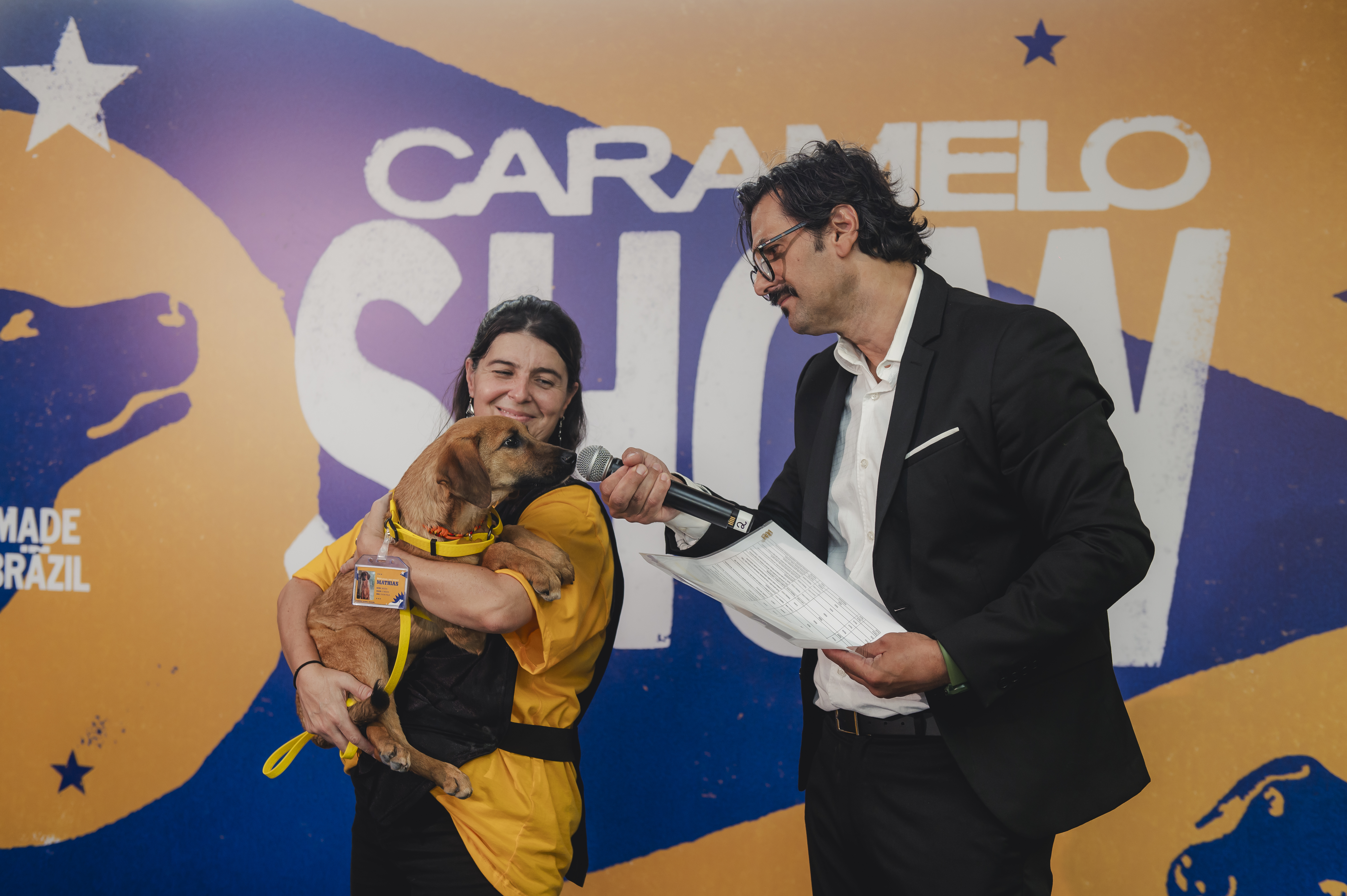 Caramelo Show com um homem de terno segurando um microfone para um cachorro caramelo no colo de uma mulher - Pedigree - Fornecedores do Assaí Atacadista.jpg