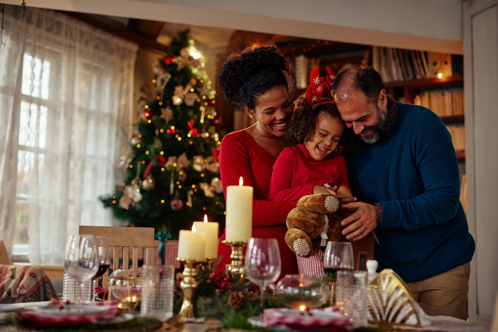 casal com uma mulher negra e um homem branco abraçando uma criança perto da mesa com a ceia de Natal - compras de Natal - Assaí Atacadista
