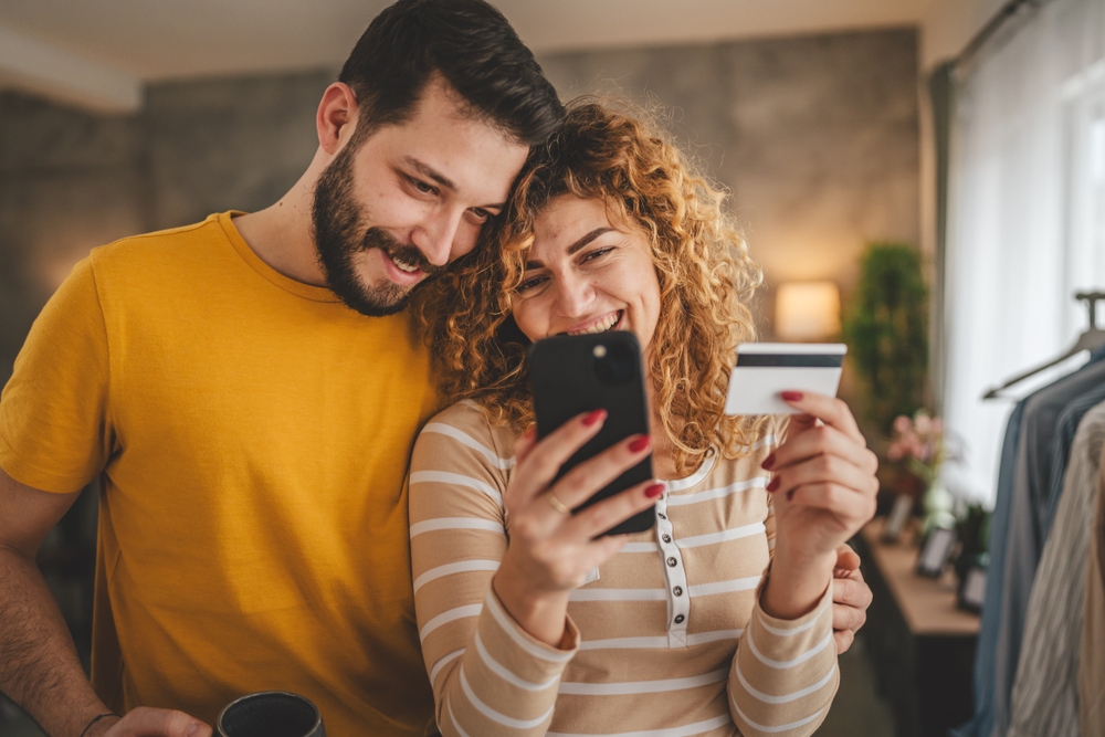 casal de homem e mulher brancos felizes abraçados segurando um celular e um cartão de crédito - Assaí Atacadista - FAQ Assaí