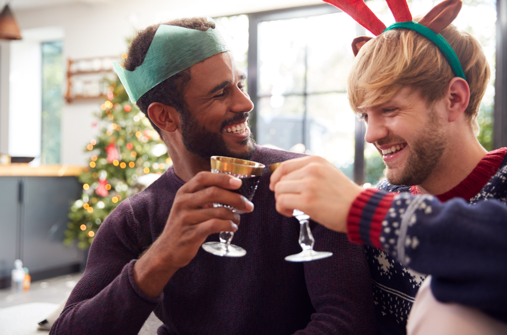 casal homoafetivo com um jovem negro e outro branco felizes brindando com uma taça e uma árvore de Natal no fundo - compras de Natal - Assaí Atacadista