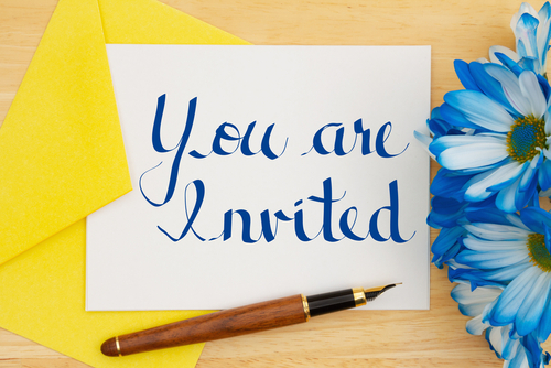 envelope amarelo com papel branco em que está escrito “você está convidado” em inglês com uma caneta de caligrafia e uma flor azul do lado - Assaí Atacadista