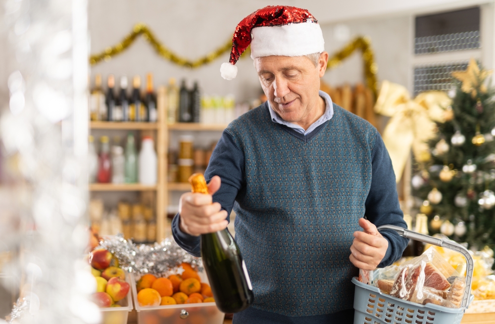 homem branco idoso com uma touca vermelha de Natal olhando para uma garrafa de champanhe em sua mão - mesa posta no Natal - Assaí Atacadista