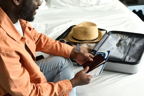 homem negro sentado na cama segurando documentos ao lado de uma mala de viagem aberta - feriado de Páscoa - Assaí Atacadista