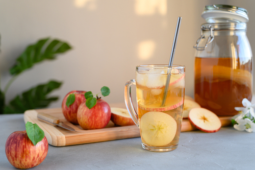 kombucha de maçã e canela em um copo com uma jarra no fundo e maçãs em uma tábua - Assaí Atacadista