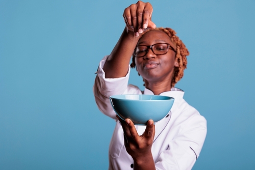 moça negra jovem com roupa de chef jogando tempero em uma tigela azul - Assaí Atacadista - temperos e especiais