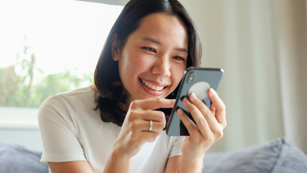 mulher asiática sorrindo enquanto vê um celular em sua mão - combate à desinformação - Assaí Atacadista
