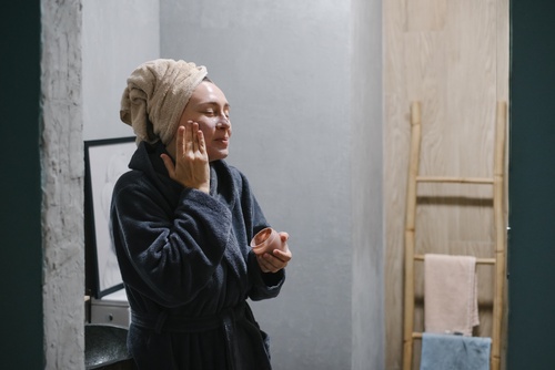 mulher branca de roupão e toalha no cabelo sorrindo e passando creme no rosto - autocuidado acessível - Assaí Atacadista