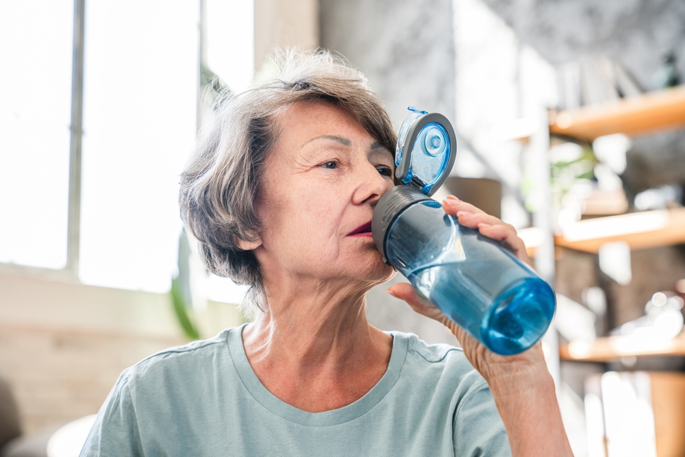 mulher branca idosa tomando água em uma garrafa azul - beleza e aiimentação - Assaí Atacadista