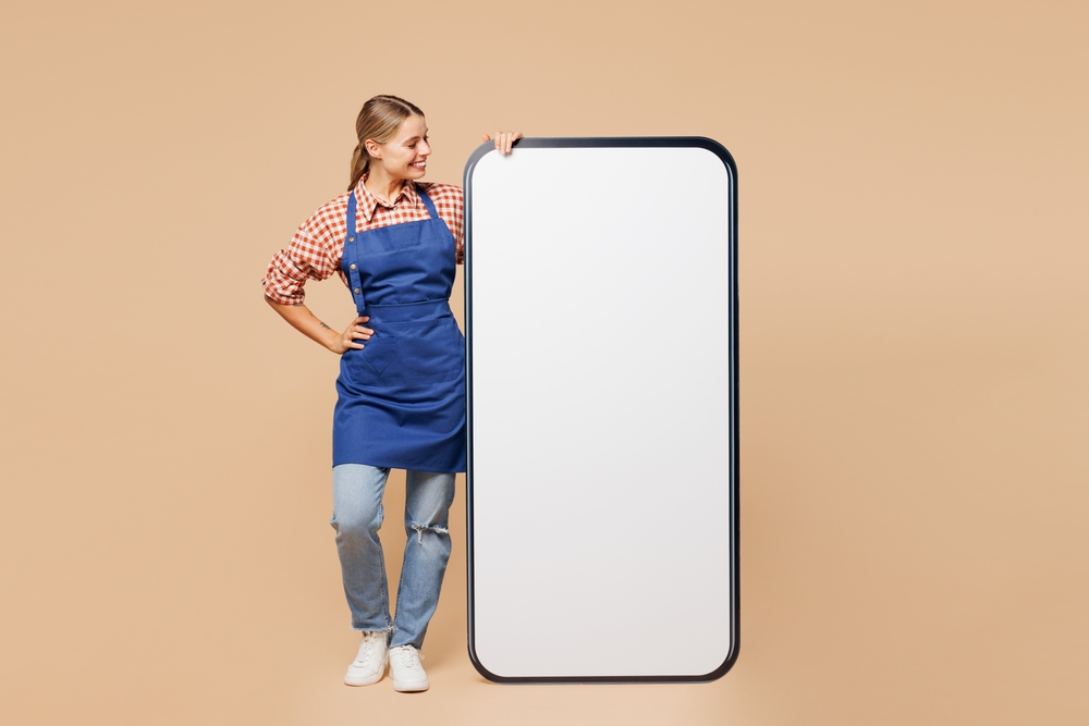 mulher branca jovem com avental sorrindo ao lado de um mockup de celular em branco - mercadinho - Assaí Atacadista