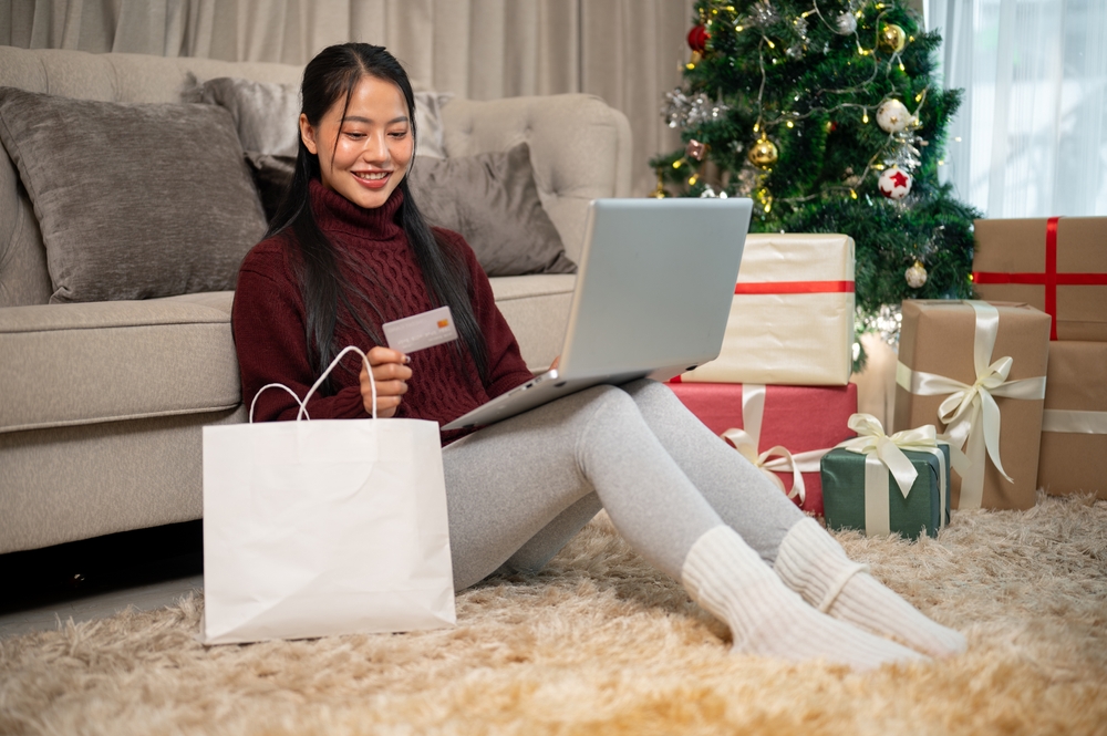 mulher jovem asiática sentada no chão com o notebook no colo e a cartão de crédito nas mãos acompanhada de presentes e uma árvore de Natal no fundo - Assaí Atacadista - delivery natalino