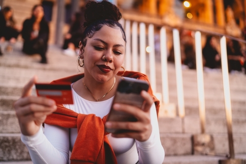 mulher negra sentada em uma escada segurando um cartão de crédito e um celular nas mãos - Assaí Atacadista - Cartão Assaí