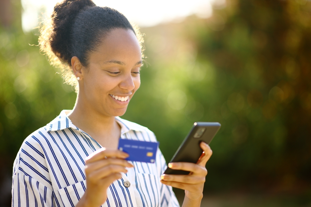 mulher negra sorrindo segurando um celular e um cartão de crédito - lanchonete em casa - Assaí Atacadista