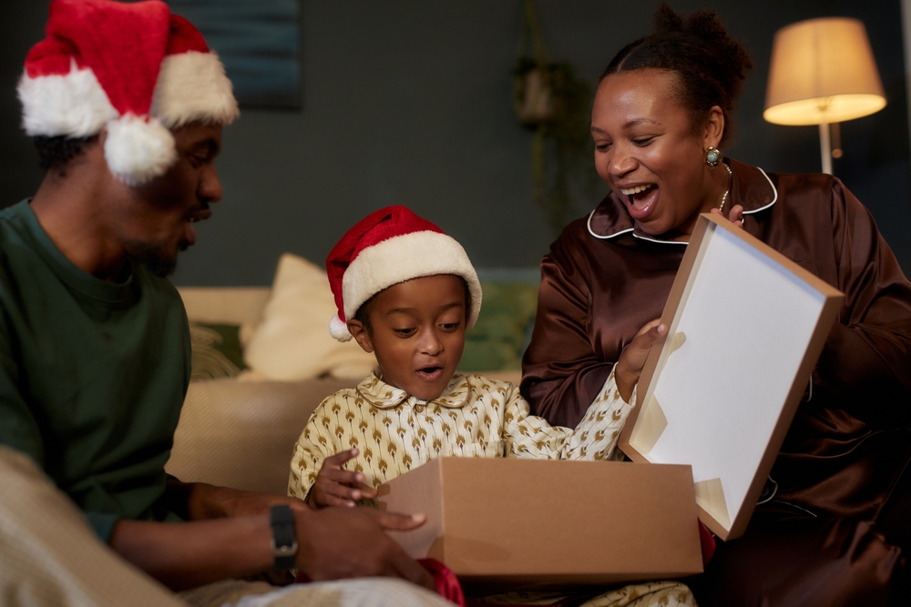 pai, mãe e criança negra felizes e surpresos enquanto abrem caixa de presente de Natal - Assaí Atacadista - atrações de Natal