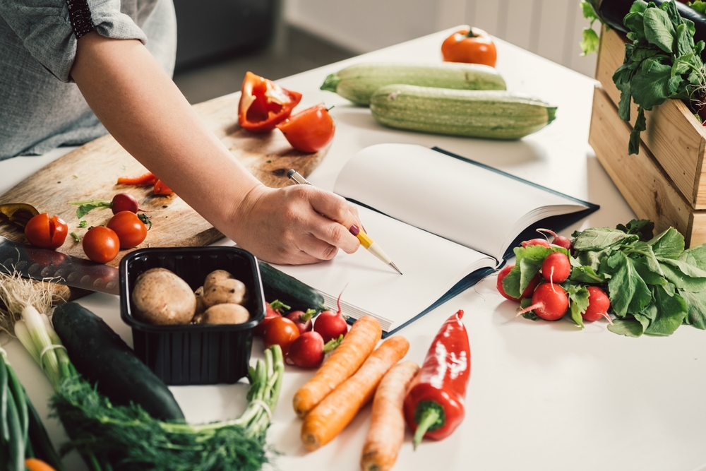 pessoa escrevendo em uma mesa de cozinha cheia de verduras e outros ingredientes - lanchonete em casa - Assaí Atacadista