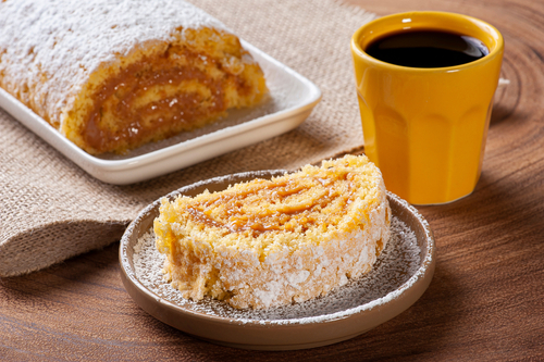 prato com rocambole de doce de leite com uma fatia cortada e uma xícara amarela de café - petiscos fáceis - Assaí Atacadista
