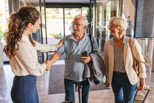 recepcionista comprimentando um homem branco idoso e uma mulher branca idosa no hall de entrada de um prédio - acomodações de Carnaval - Assaí Atacadista - Imagem: Shutterstock
