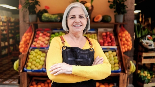senhora empreendedora feliz com os braços cruzados em frente a uma bancada de frutas - Dia do Consumidor - Assaí Atacadista