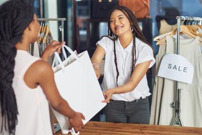 moça sorrindo entregando uma sacola para uma cliente em uma loja de roupas - Assaí Atacadista - atendimento ao cliente