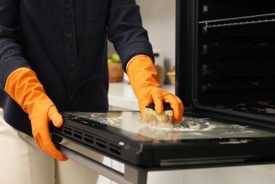 pessoa com luvas de limpeza laranja esfregando um fogão com uma esponja - limpeza da cozinha - Assaí Atacadista