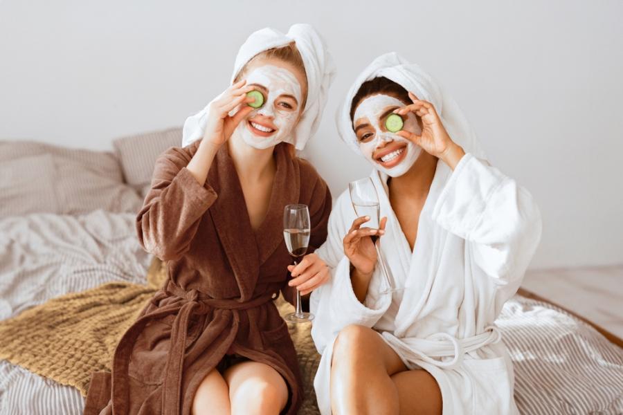 duas mulheres felizes em roupão e máscara facial segurando um copo e uma rodela de pepino nas mãos - spa em casa - Assaí Atacadista