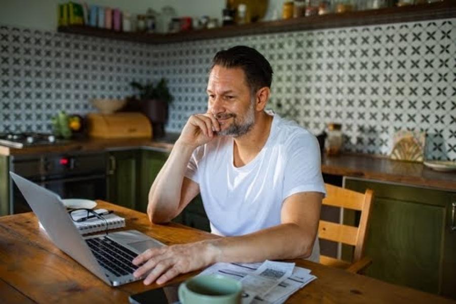 homem branco da terceira idade sentado na cozinha sorrindo enquanto olha para um notebook - Assaí Atacadista - rotina pós-Carnaval - Imagem: Shutterstock