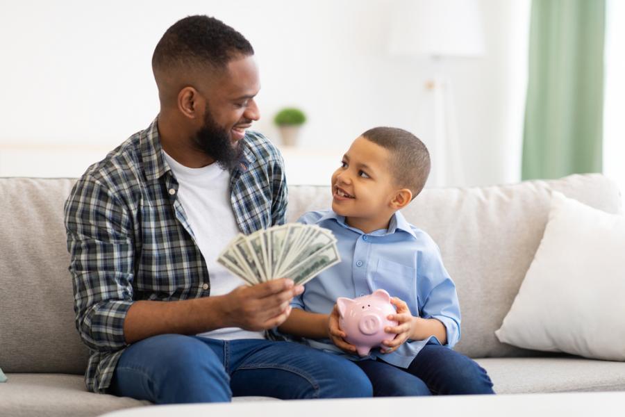 homem negro segurando notas de dinheiro nas mãos ao lado de um menino negro feliz segurando um cofre de porquinho - poupar dinheiro com filhos - Assaí Atacadista