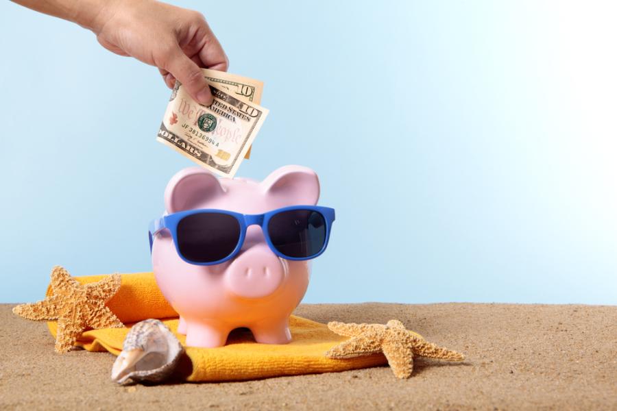 mão branca colocando uma nota de dolar em um cofre no formato de porquinho com óculos de sol - renda extra no verão - Assaí Atacadista