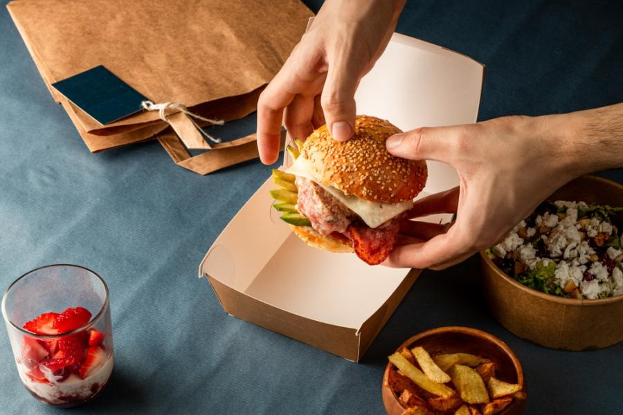 mãos brancas segurando um hamburguer com outros alimentos na mesa - lanchonete em casa - Assaí Atacadista