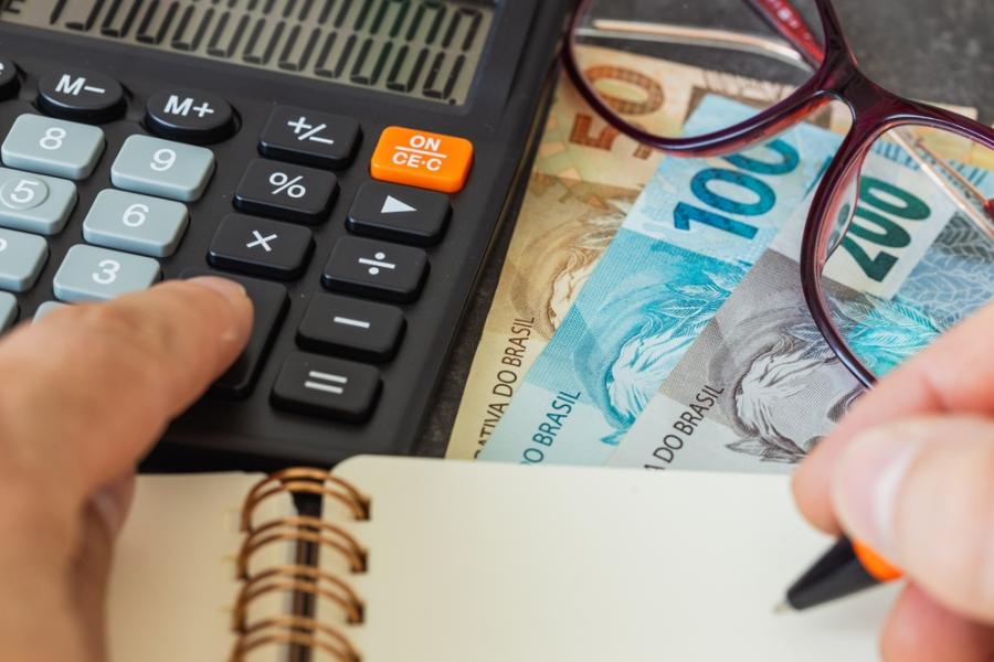 mãos clicando em uma calculadora e escrevendo em um caderno com notas de dinheiro e um óculos de grau na mesa - dicas de economia no verão - Assaí Atacadista