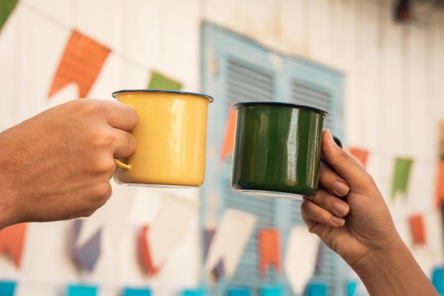 mãos de duas pessoas brindando xicaras com bandeirinhas de são joão no fundo - bebidas de festa junina - Assaí Atacadista