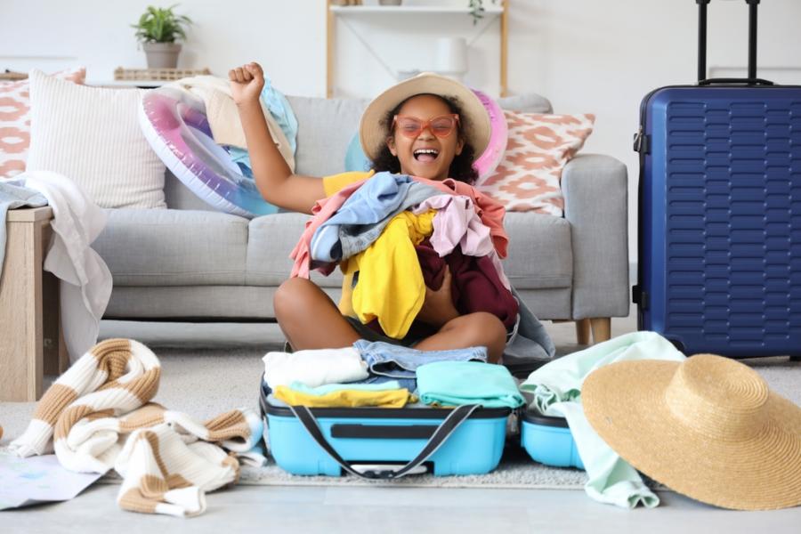 menina negra feliz segurando roupas em uma mala de viagem - destinos de viagem verão - Assaí Atacadista