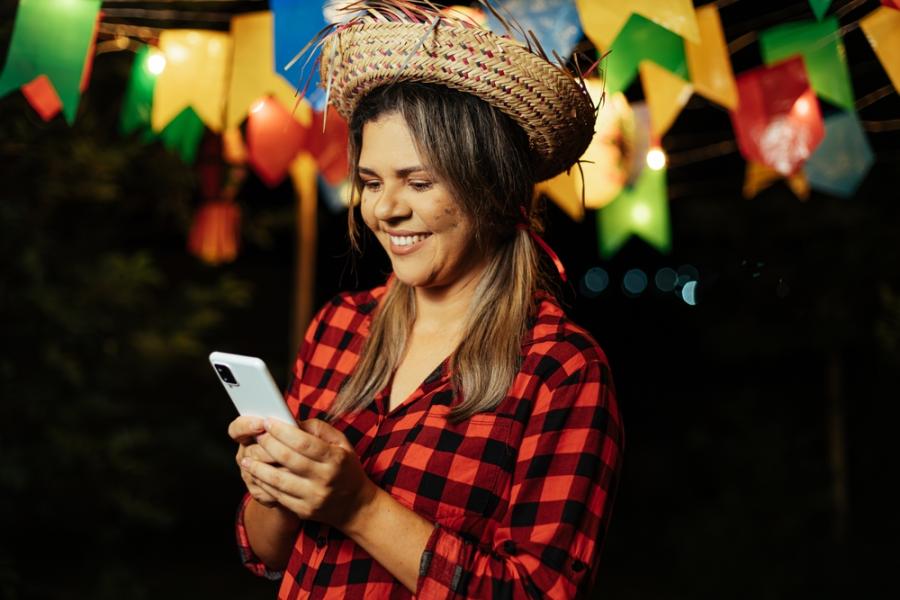 mulher branca com trajes de festa junina segurando e olhando para o celular sorrindo - curiosidades sobre o São João - Assaí Atacadista