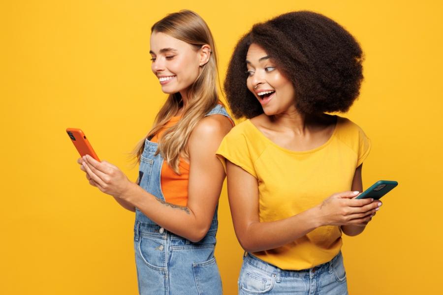 mulher branca e mulher negra felizes segurando um celular em um fundo amarelo-App Meu Assaí-Assaí Atacadista