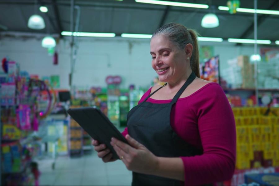 mulher branca empreendedora sorrindo olhando para um tablet no meio de um mercadinho - Assaí Atacadista