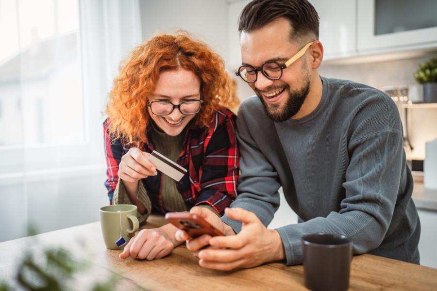 mulher e homem brancos apoiados em uma bancada olhando para o celular felizes - Dia do Consumidor - Assaí Atacadista - Imagem: Shutterstock