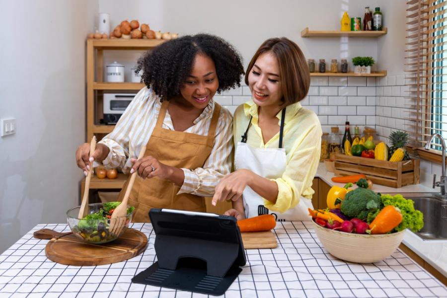mulher preta e mulher asiática preparando uma receita de salada felizes na cozinha - Assaí Atacadista