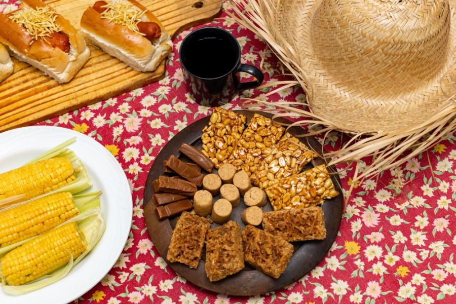 pratos com comidas típicas de São João e um chapéu de palha em uma toalha floral - Assaí Atacadista