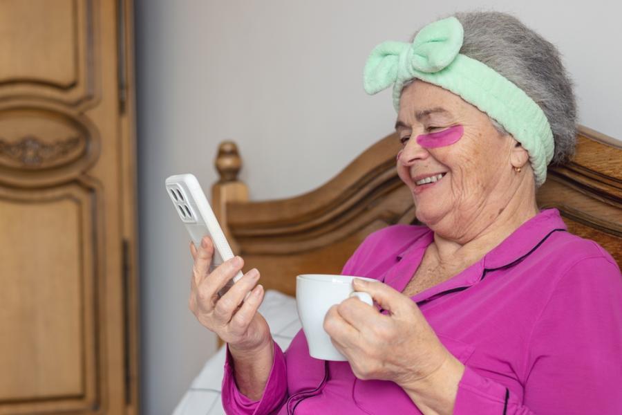 senhora branca segurando um celular e uma xícara nas mãos sentada na cama com uma mascara nas olheiras e uma faixa no cabelo - produtos de autocuidado - Assaí Atacadista