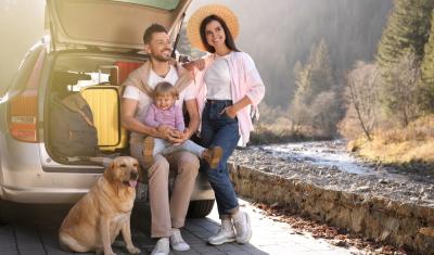 carro com porta malas aberto com um homem branco sentado com uma bebê no colo e uma mulher branca em pé do lado e um cachorro labrador no chão - viagem de páscoa - Assaí Atacadista