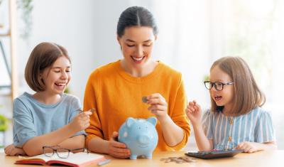 duas crianças e uma mulher olhando pra um cofre de porquinho em uma mesa-educação financeira infantil-Assaí Atacadista