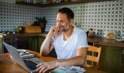 homem branco da terceira idade sentado na cozinha sorrindo enquanto olha para um notebook - Assaí Atacadista - rotina pós-Carnaval - Imagem: Shutterstock