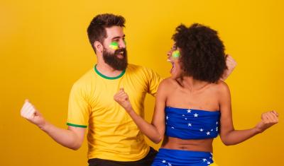 homem e mulher com as roupas da bandeira do Brasil felizes olhando um para o outro em um fundo amarelo-casal torcedor-Assaí Atacadista