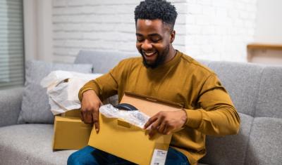 homem negro feliz sentado no sofá abrindo uma caixa - clube de assinatura - Assaí Atacadista