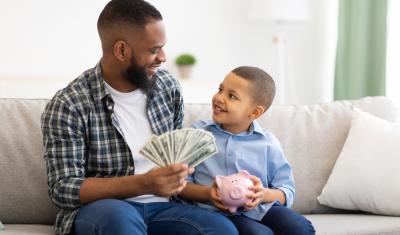 homem negro segurando notas de dinheiro nas mãos ao lado de um menino negro feliz segurando um cofre de porquinho - poupar dinheiro com filhos - Assaí Atacadista