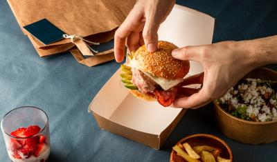 mãos brancas segurando um hamburguer com outros alimentos na mesa - lanchonete em casa - Assaí Atacadista