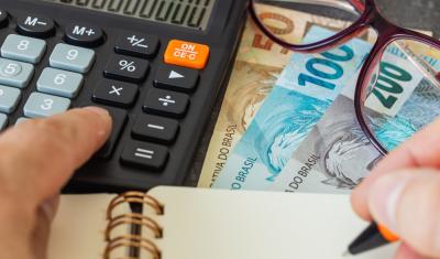 mãos clicando em uma calculadora e escrevendo em um caderno com notas de dinheiro e um óculos de grau na mesa - dicas de economia no verão - Assaí Atacadista