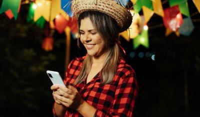 mulher branca com trajes de festa junina segurando e olhando para o celular sorrindo - curiosidades sobre o São João - Assaí Atacadista