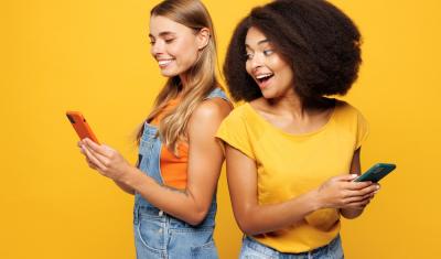 mulher branca e mulher negra felizes segurando um celular em um fundo amarelo-App Meu Assaí-Assaí Atacadista