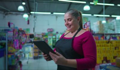 mulher branca empreendedora sorrindo olhando para um tablet no meio de um mercadinho - Assaí Atacadista