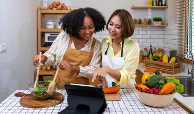 mulher preta e mulher asiática preparando uma receita de salada felizes na cozinha - Assaí Atacadista